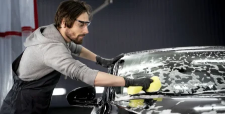 Майстер автосервісу миє лобове скло чорного автомобіля спеціальною активною піною та жовтою губкою.